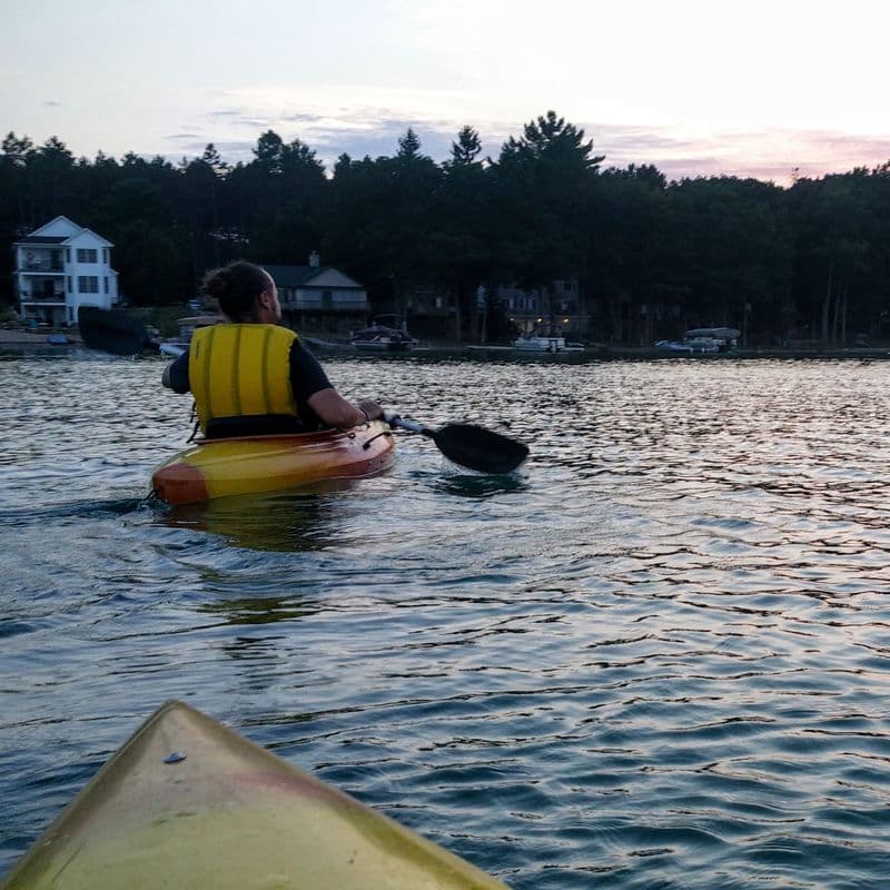A kayaker paddling on calm water at golden hour, with a wooded shoreline of docks and cottages behind.